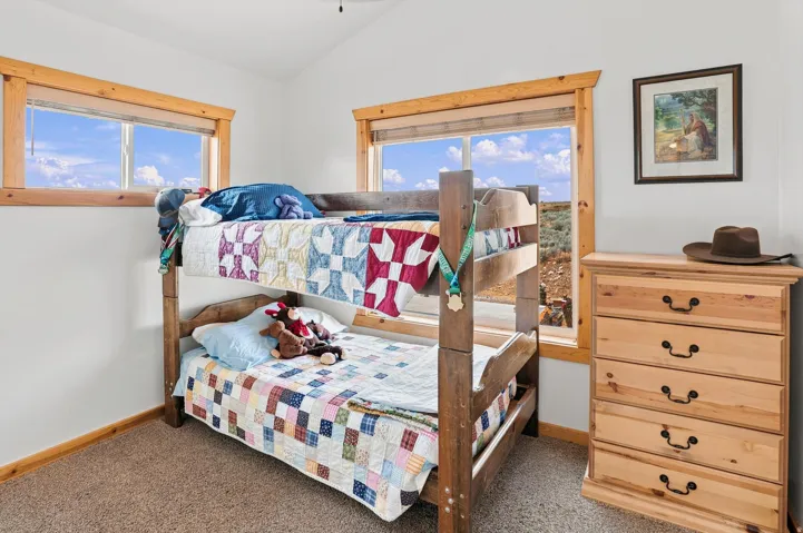 Interior room featuring a wood-frame bunk bed, natural wood trim around windows and baseboards, two windows with outdoor scenery, and light-colored carpet flooring