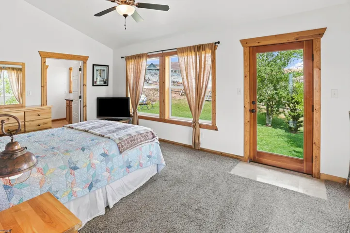 Primary Suite Vaulted ceiling with a ceiling fan, three pane windows with wood trim, a full-glass door with wood trim, and grey carpet flooring