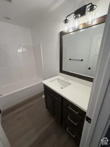 Bathroom featuring a white tub/shower combination, wood-finish flooring, a dark wood-finish vanity with an integrated sink, a white countertop, and a framed mirror with a three-light fixture