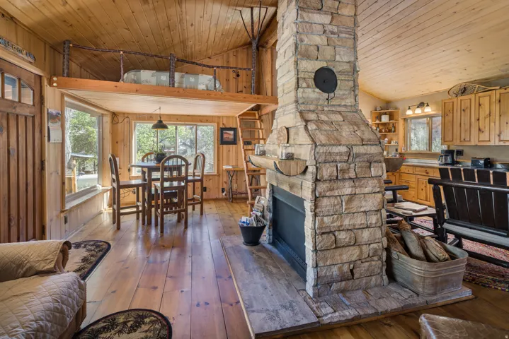Stone-clad fireplace with hearth, wood-finish flooring, wood-paneled walls and ceiling, built-in kitchen cabinetry, and an upper loft area