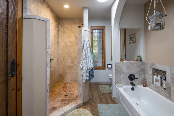 Spacious bathroom featuring a walk-in shower with tile surround and floor, a built-in bathtub with tile surround and recessed shelving, wood-finish flooring, and a round skylight