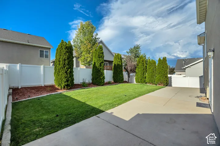 Private backyard featuring a concrete patio, manicured lawn, white vinyl fencing, and mature evergreen trees