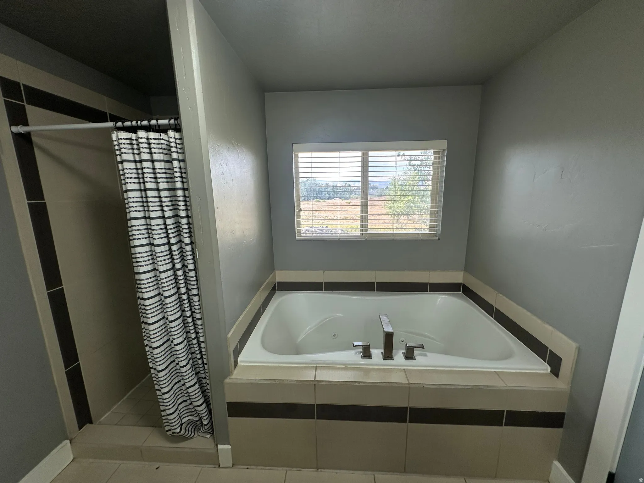 Soaking tub with a tile surround, window with horizontal blinds, and adjacent shower stall