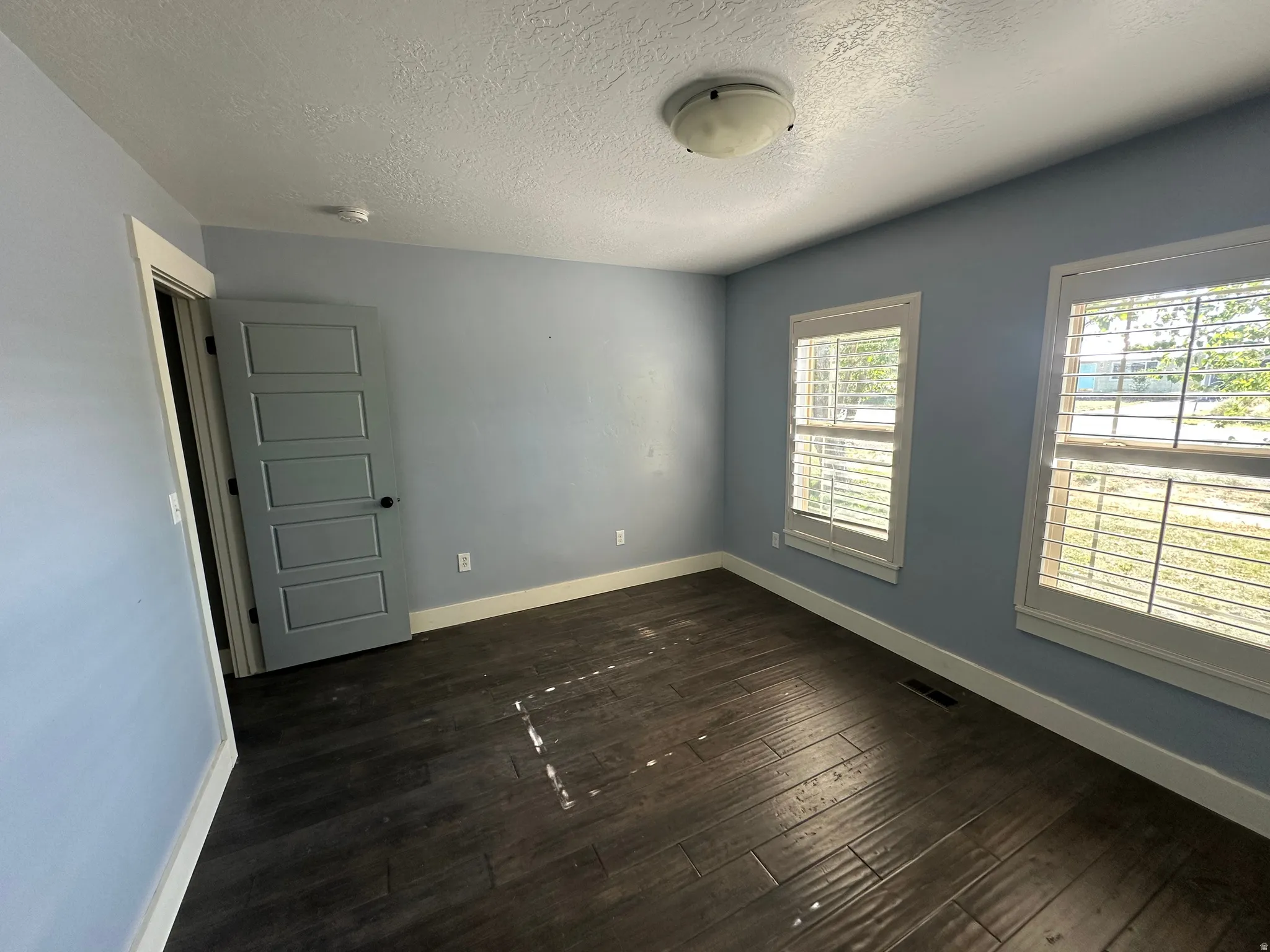 Room featuring dark wood-finish flooring, two large windows with white louvered blinds, and a five-panel interior door with a black handle
