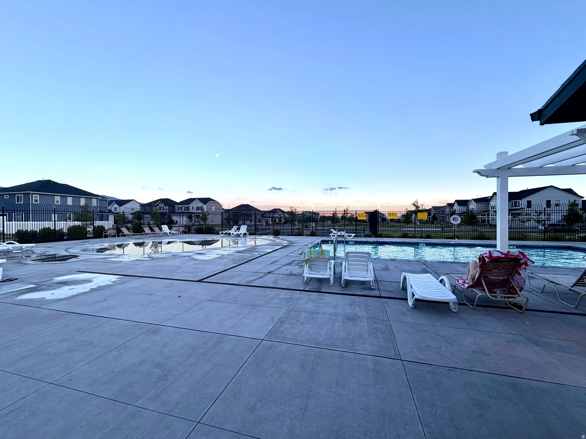 Community swimming pool with a surrounding concrete deck, black perimeter fencing, and white pergola