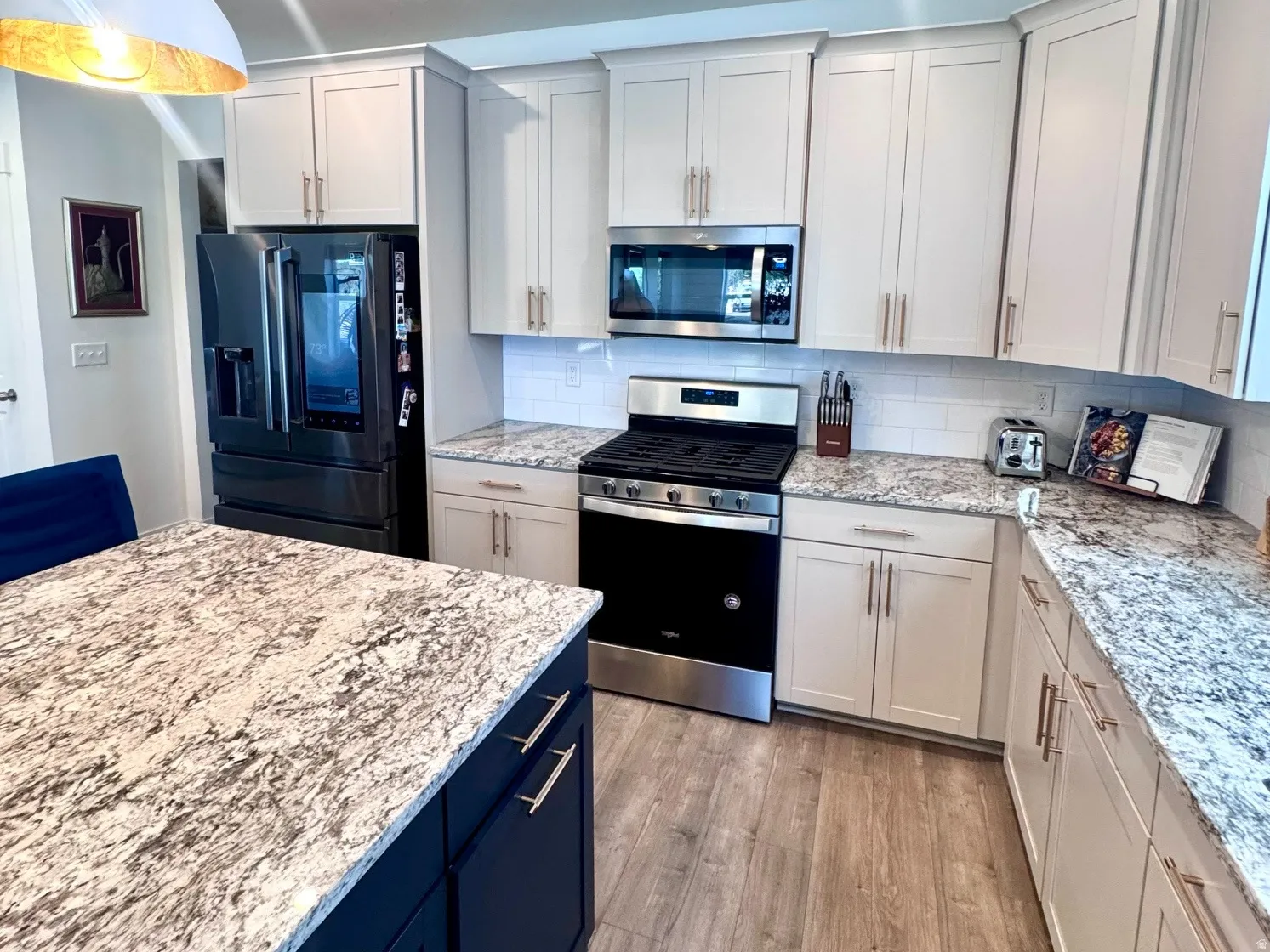 Contemporary kitchen featuring an expansive center island with dark cabinetry, light-toned perimeter cabinetry, stainless steel appliances, speckled countertops, and wood-finish flooring