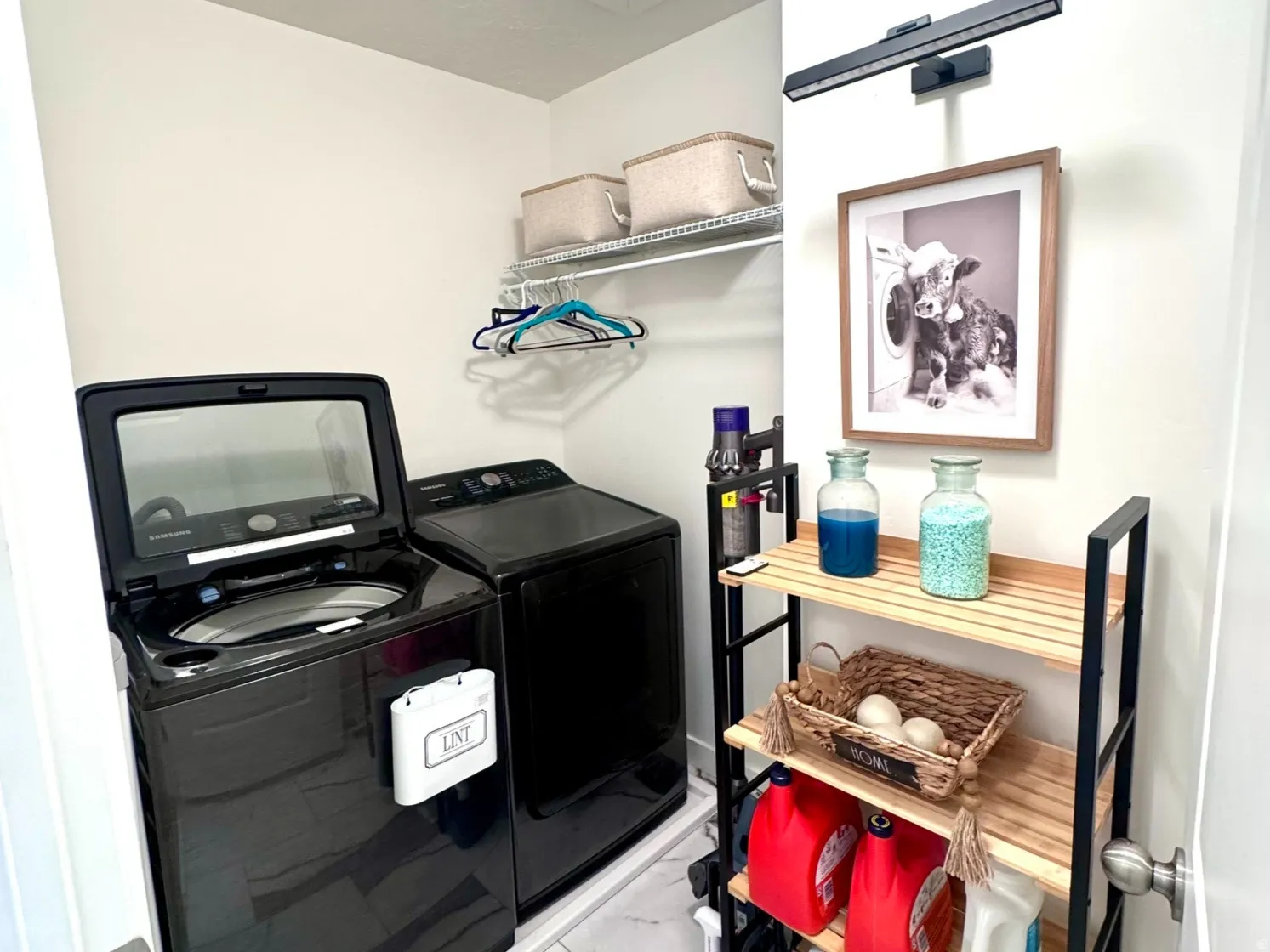 Laundry area featuring marble-finish tile flooring, a built-in wire shelf, overhead lighting, and a slatted wood-and-metal shelving unit