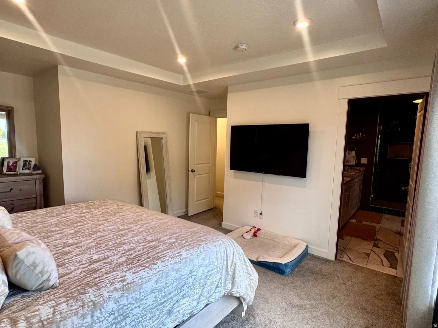 Spacious room featuring tray ceilings with recessed lighting, plush carpeting, a window providing natural light, and a door opening into an adjoining tiled space