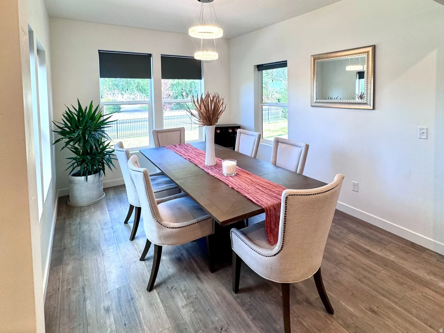 Spacious dining area featuring wood-finish flooring, multiple windows providing natural light, a modern multi-ring pendant light fixture, and an open beam ceiling