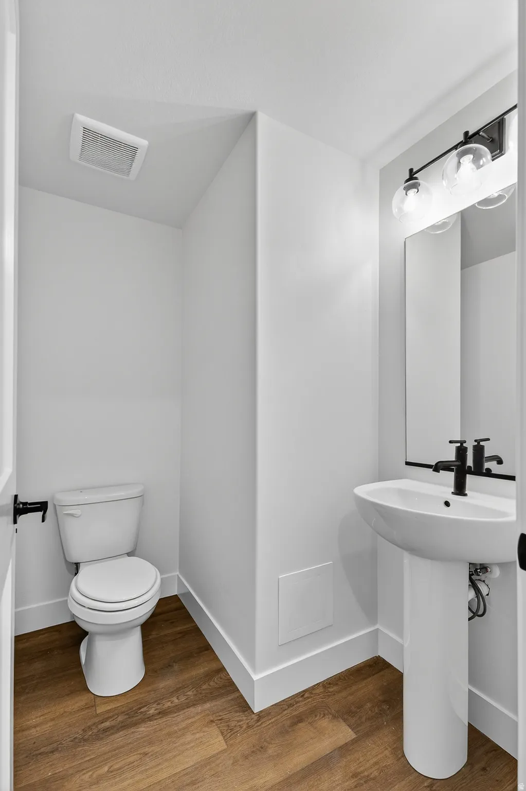 Powder room with wood-finish flooring, white pedestal sink, black wall-mounted faucet, and a three-light vanity fixture