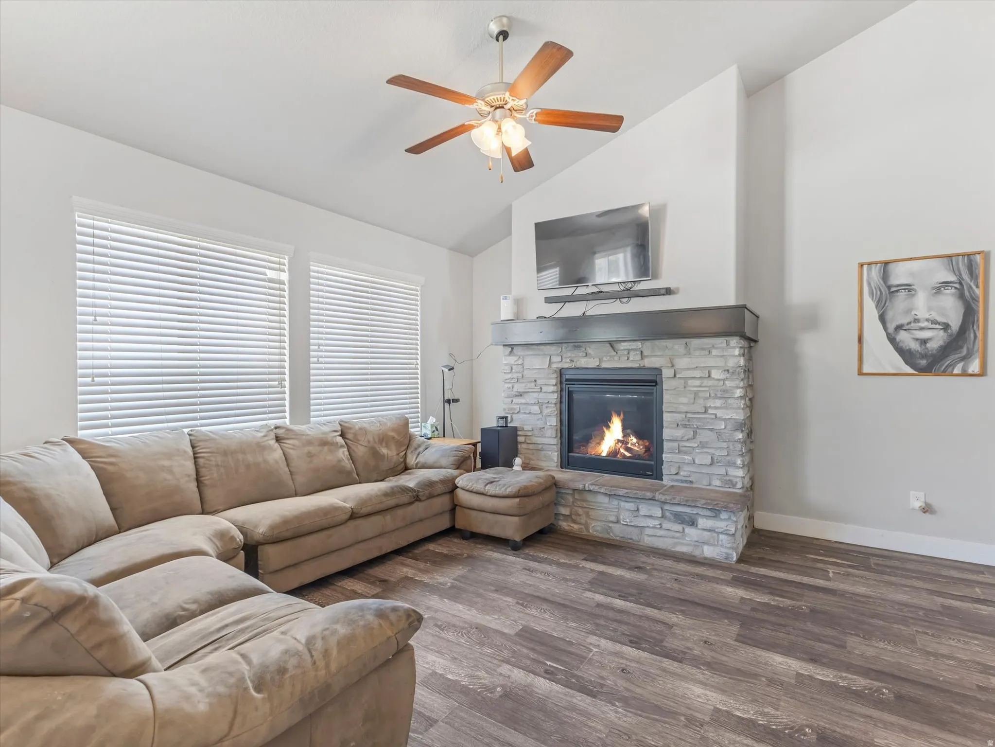 Vaulted ceiling living room featuring a stone-clad fireplace with a dark wood-finish mantel, two large windows with blinds, wood-finish flooring, and a ceiling fan