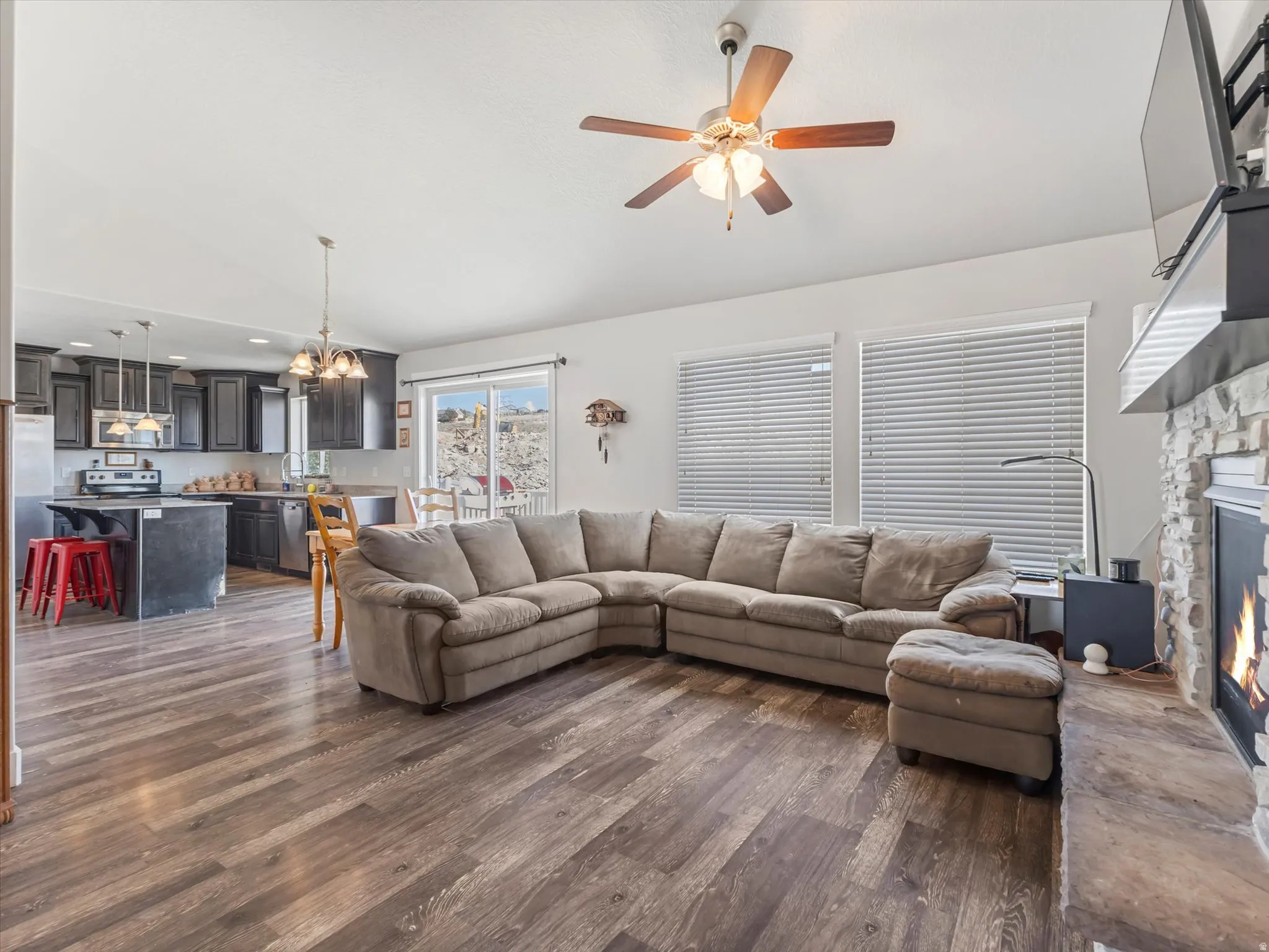 Open-concept living space featuring wood-finish flooring, a stone-clad fireplace with a hearth, and a ceiling fan
