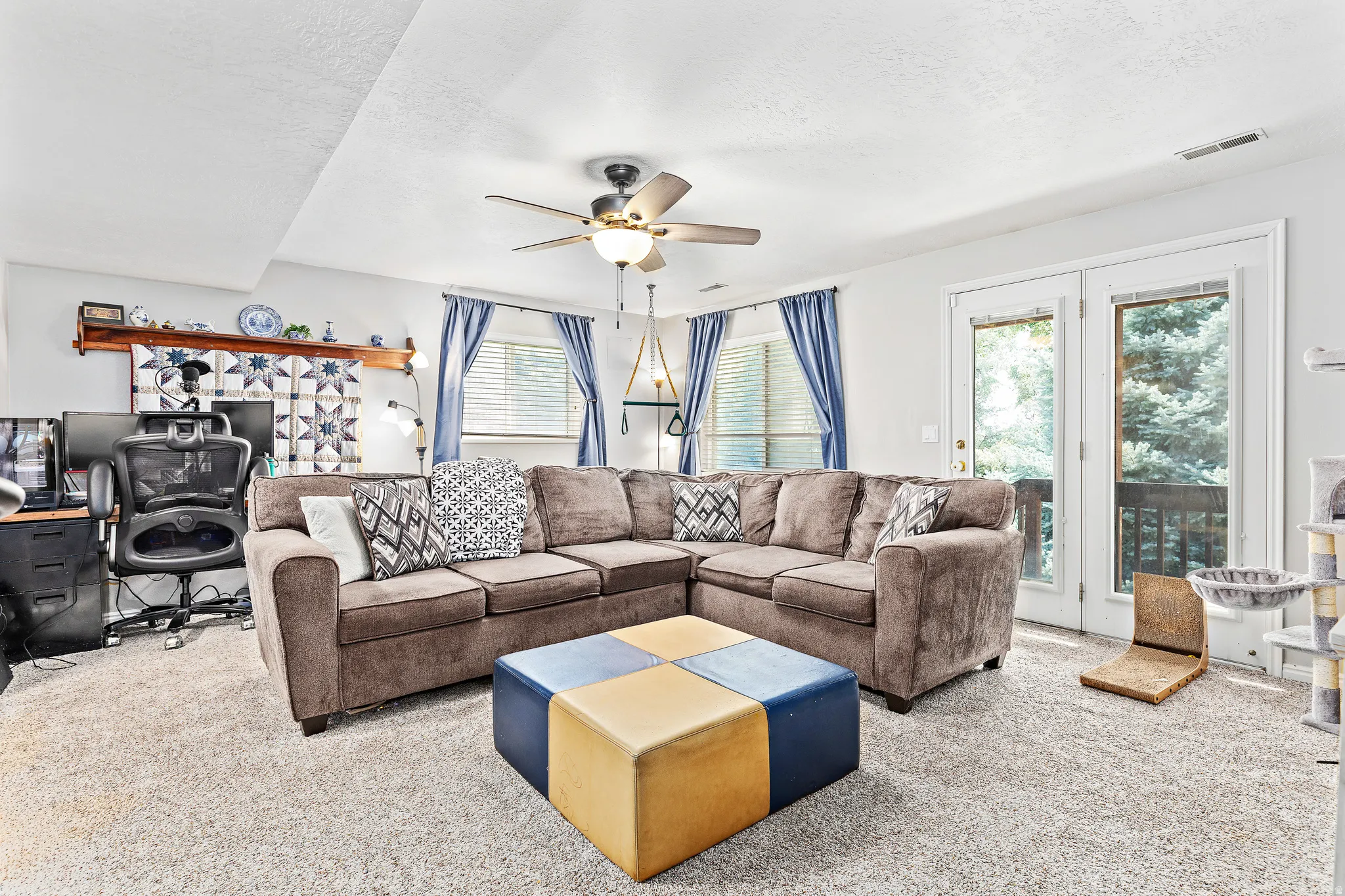 Carpeted room featuring a ceiling fan, dual windows with blinds and blue curtains, and double glass panel doors opening to an exterior balcony
