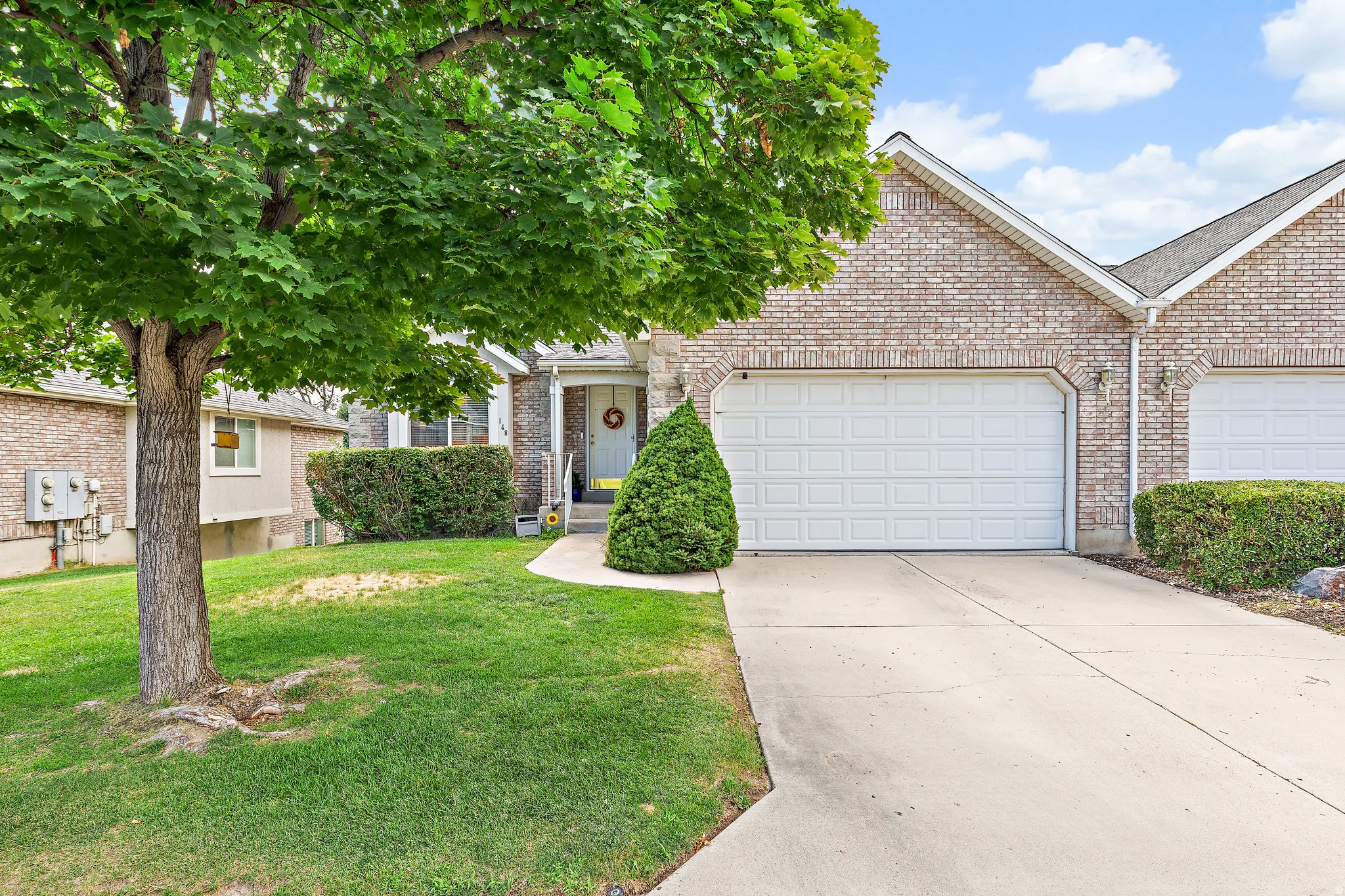 Brick exterior featuring a two-car garage with white panel doors, concrete driveway, front lawn, and mature tree