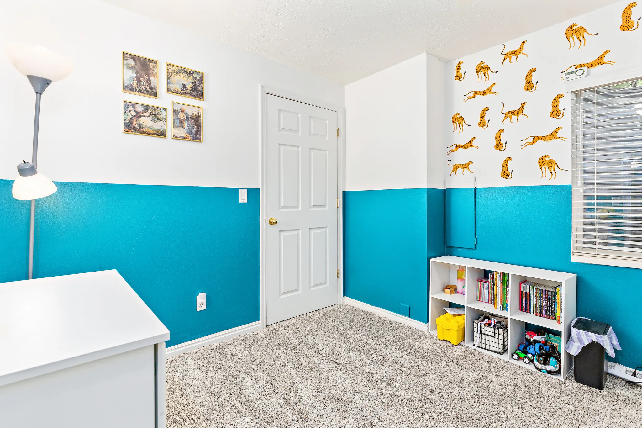Carpeted room with a two-tone wall design, featuring a white paneled door, a window with horizontal blinds, and a built-in white shelving unit