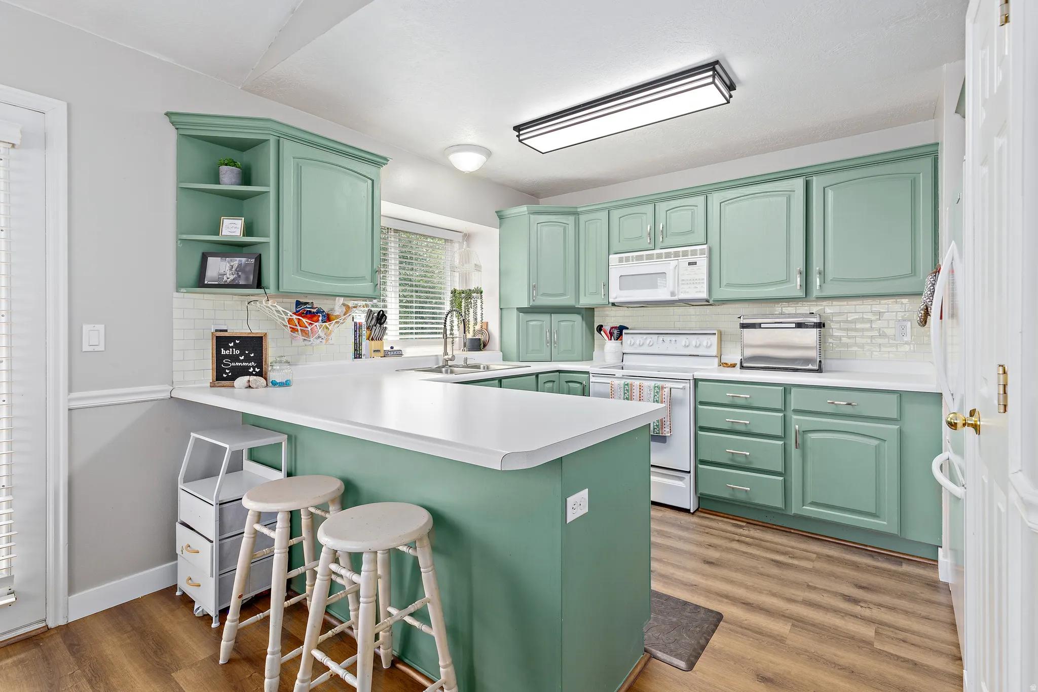 Kitchen featuring L-shaped counter space with a breakfast bar, sage-toned cabinetry, white appliances, tile backsplash, and wood-finish flooring