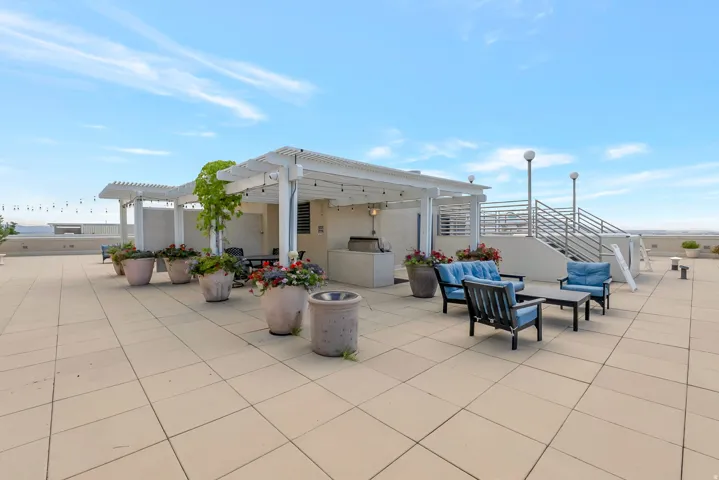 Expansive rooftop patio featuring large format tile flooring, a white pergola structure, built-in outdoor kitchen area, and string lighting