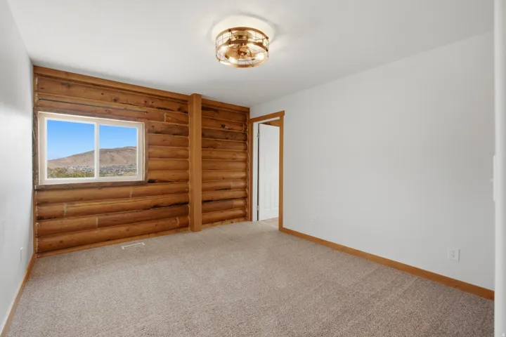 Room featuring a log-style accent wall with a large window, neutral carpeting, white painted walls, and a contemporary ceiling light fixture