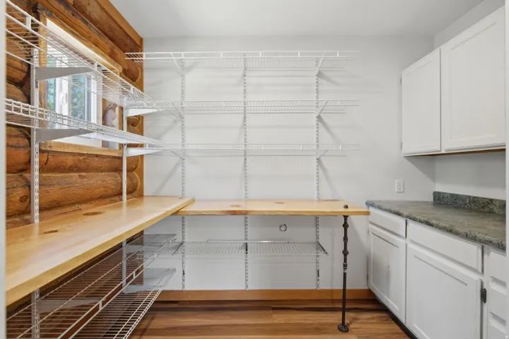 Spacious utility area featuring exposed log wall, light wood-finish flooring, white wire shelving, a natural wood countertop, and white shaker-style cabinetry with a dark countertop