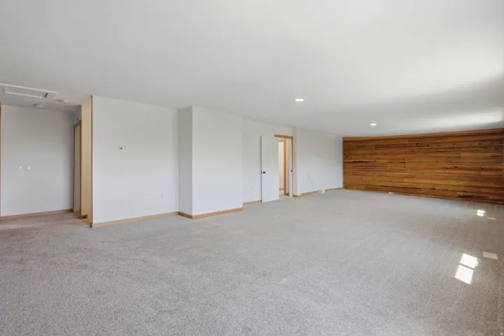 Spacious room featuring light gray carpeting, crisp white walls, recessed ceiling lighting, a distinctive wood panel accent wall, and wood-trim doorways