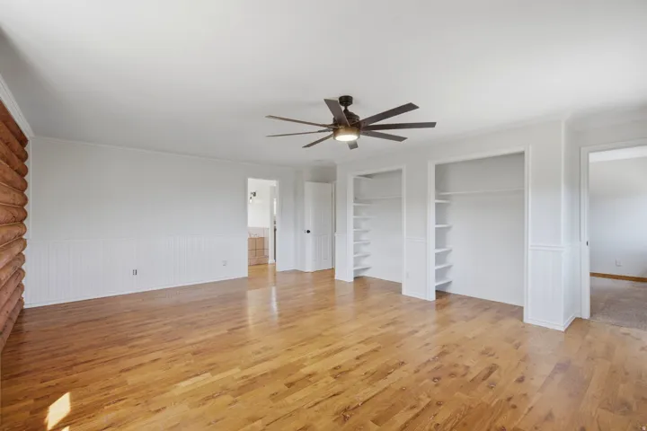 Spacious room featuring wood-finish flooring, a log-style accent wall, white wainscoting, and a multi-blade ceiling fan