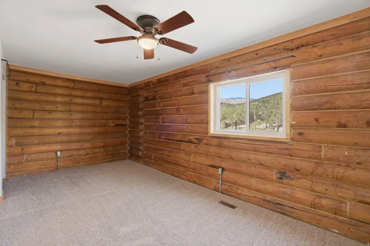 Room featuring wood-finish log walls, a ceiling fan with light fixture, light-colored carpet flooring, and a window with a mountain and tree landscape visible