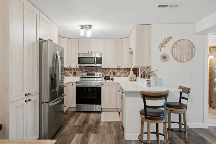 Kitchen featuring wood-finish flooring, light-toned cabinetry, stainless steel appliances, a multi-tone tile backsplash, and a white countertop with an integrated breakfast bar