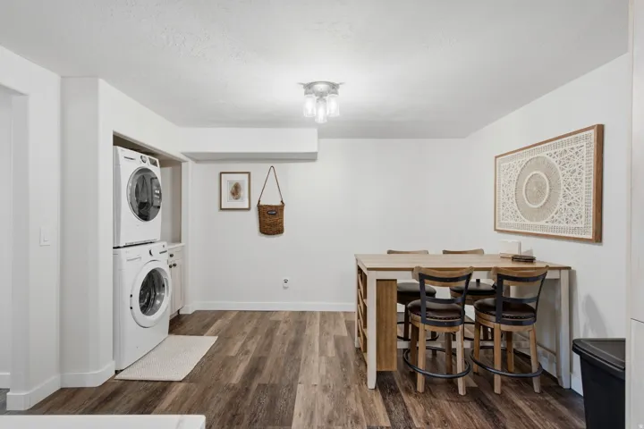 Laundry area with a stacked washer and dryer, adjacent to a dining space featuring wood-finish flooring and white walls