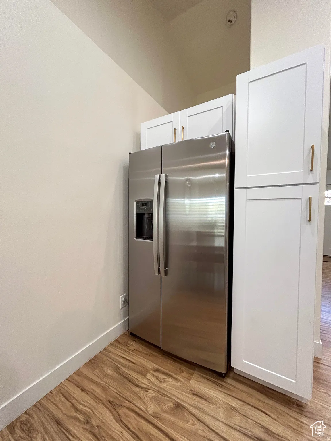 Stainless steel side-by-side refrigerator, white shaker-style cabinetry with gold-tone hardware, wood-finish flooring, and crisp white baseboards