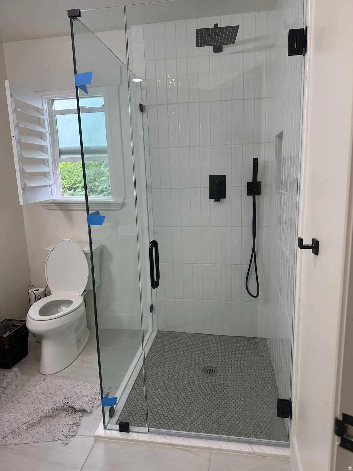 Modern glass-enclosed shower featuring vertically stacked white subway tile, a dark penny tile floor, a black rainfall showerhead, and a black handheld shower sprayer