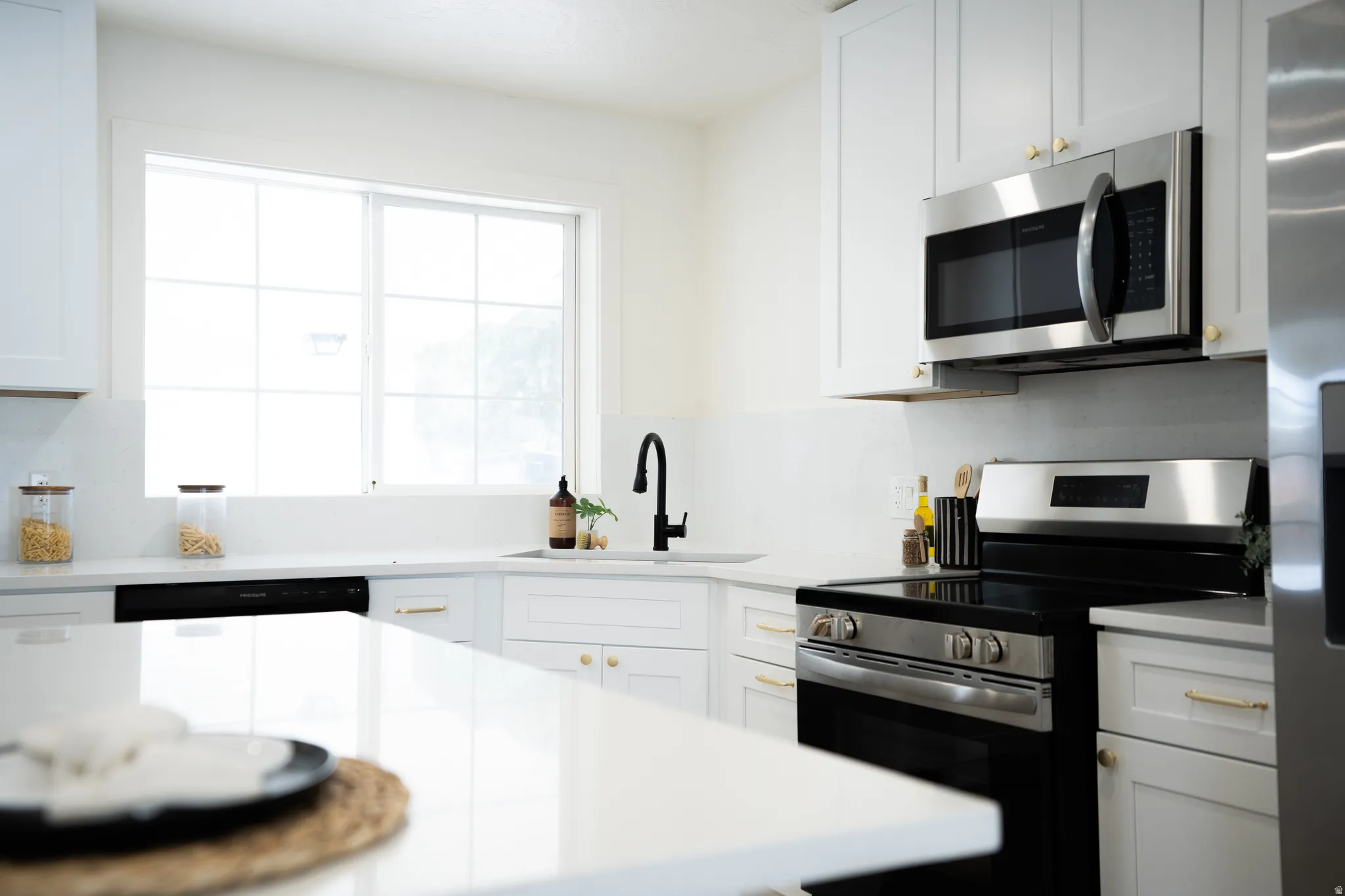 Modern kitchen featuring white shaker cabinetry, stainless steel appliances, a white solid-surface countertop, an undermount sink with a matte black gooseneck faucet, and a large window with grid panes