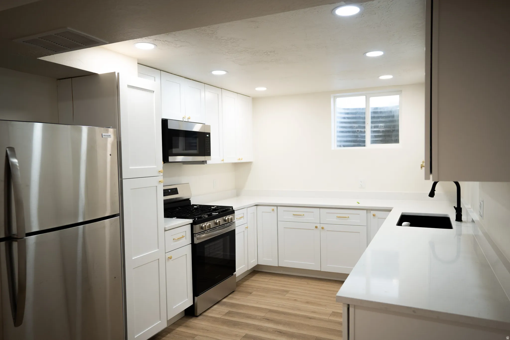Modern kitchen featuring stainless steel appliances, white shaker cabinetry with gold hardware, light wood-finish flooring, recessed lighting, and a window with a window well