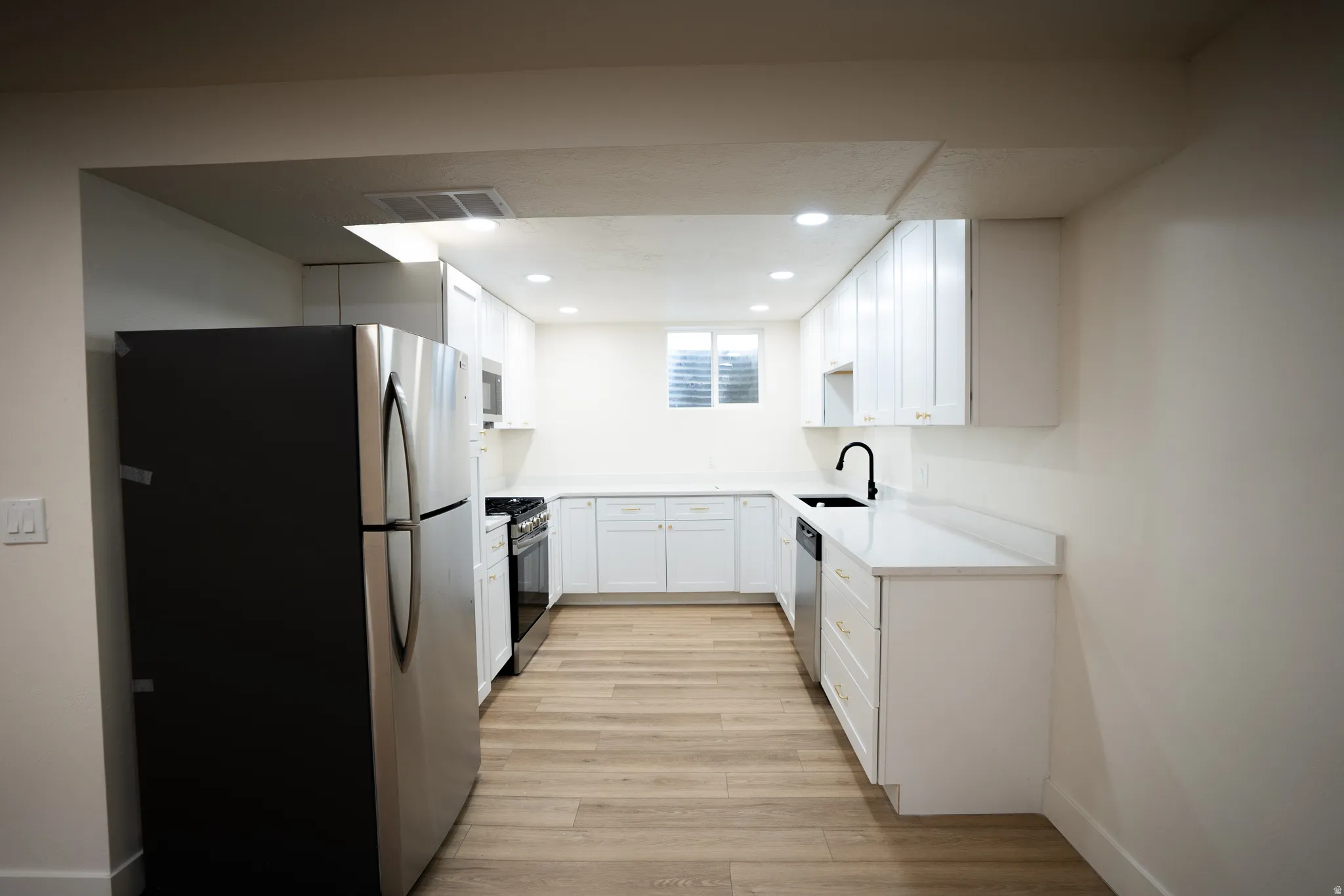 Galley kitchen featuring wood-finish flooring, recessed lighting, white cabinetry with gold-tone hardware, and stainless steel appliances