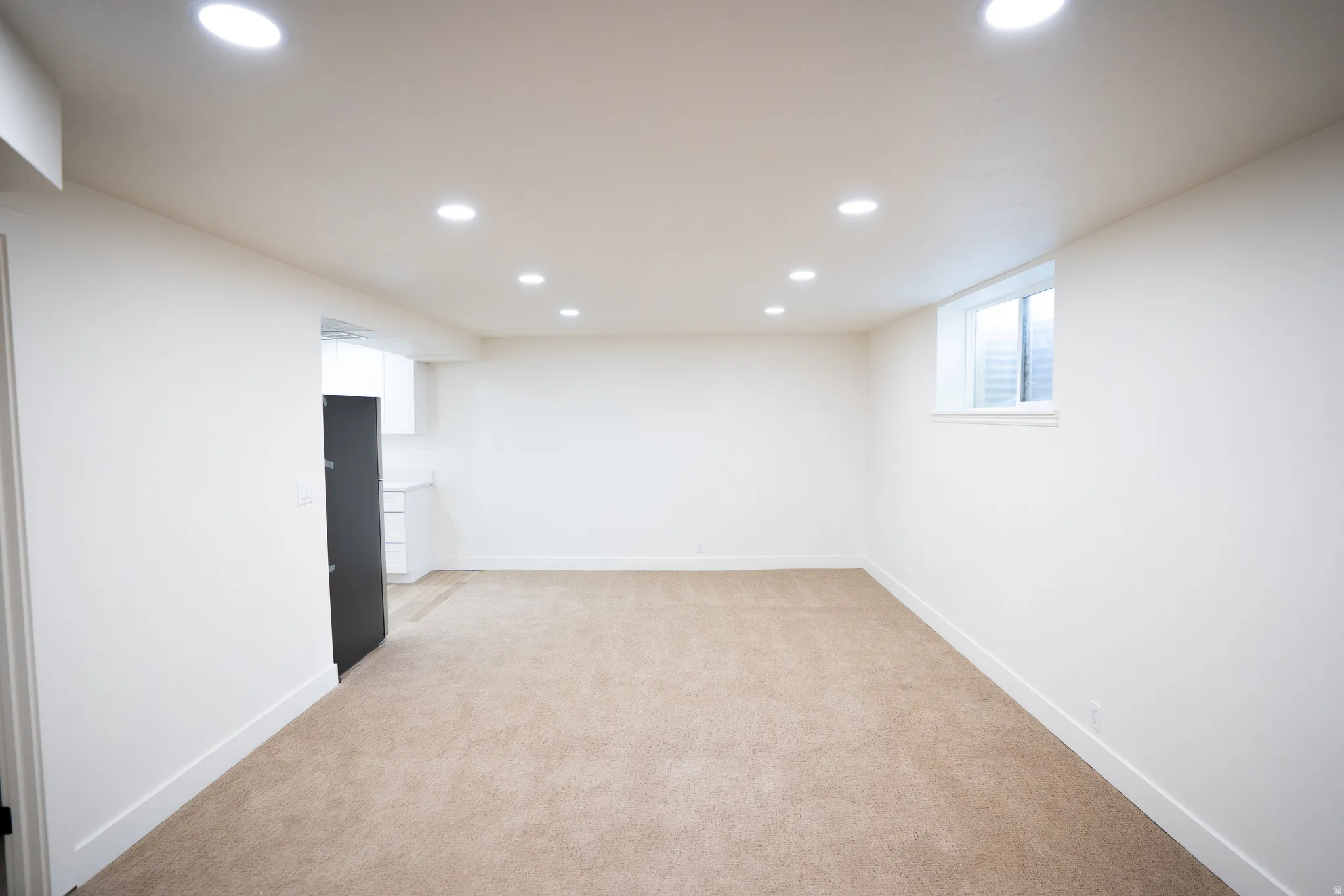 Open-concept living area featuring recessed lighting, a neutral carpet, a large egress window, and a partial view of a kitchen area with white cabinetry and a dark refrigerator