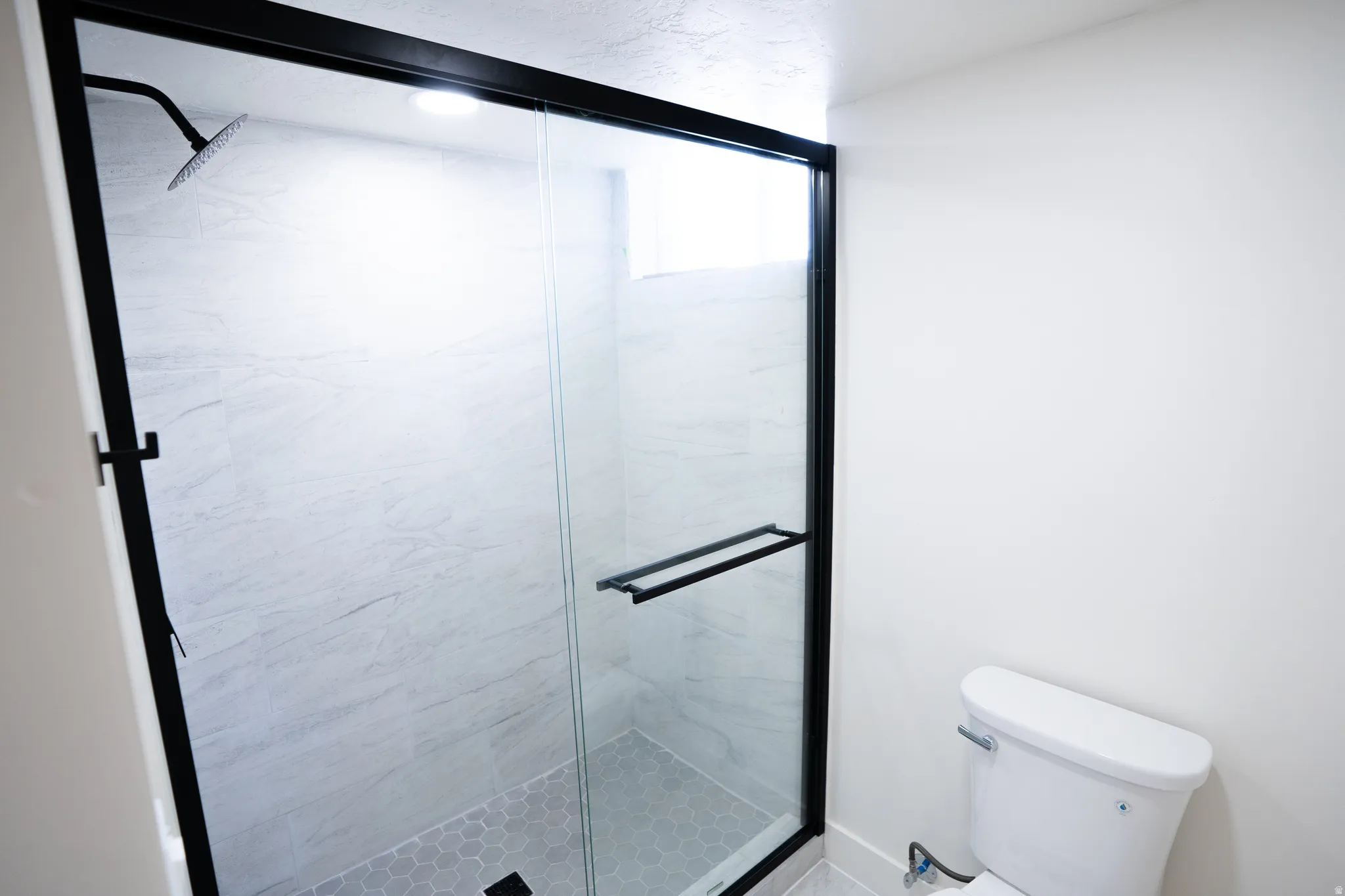 Walk-in shower featuring a frameless glass enclosure with black trim, a rain shower head, light-toned large format tile walls, and hexagonal mosaic floor tiling