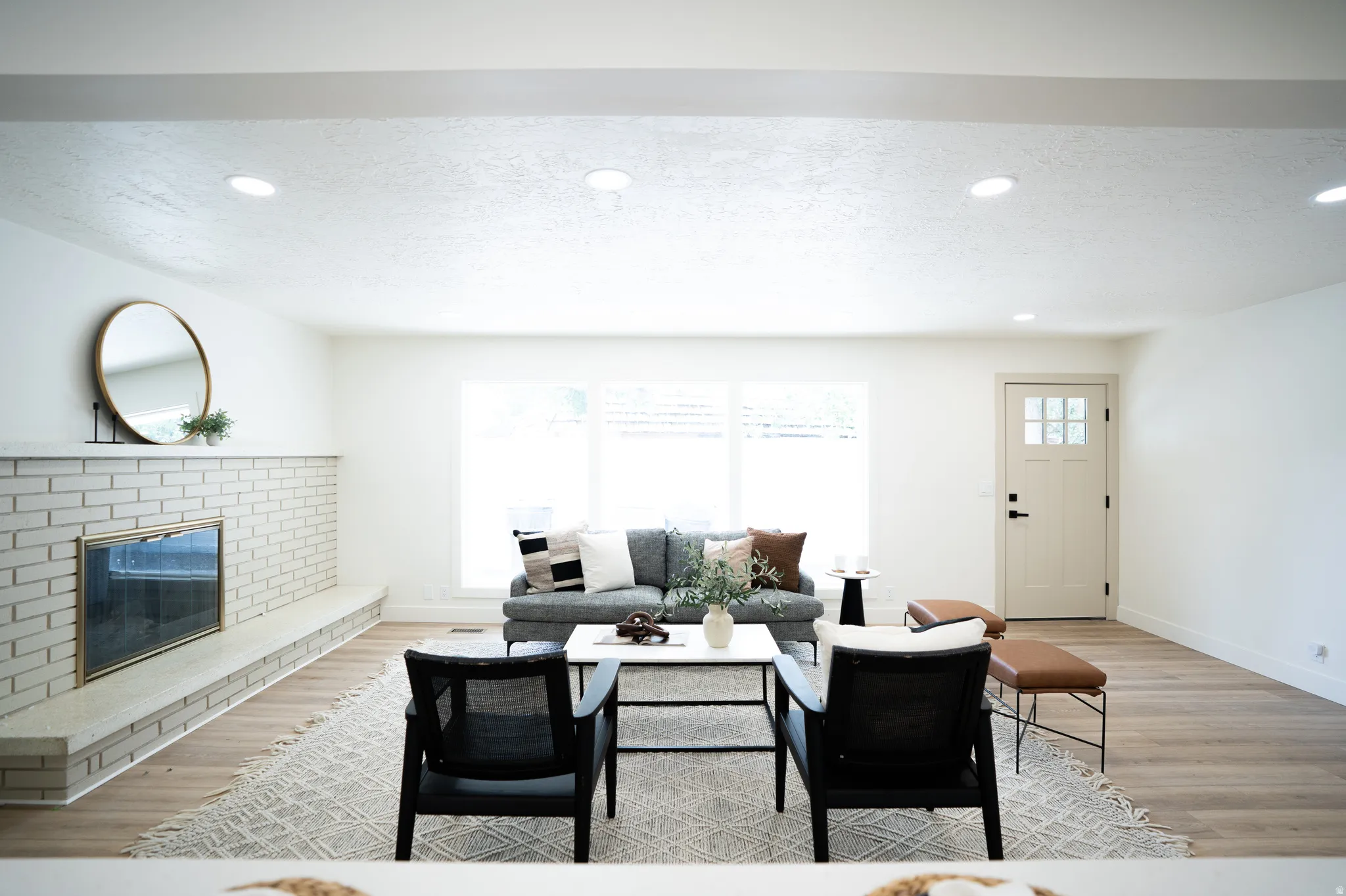Spacious living area featuring light wood-finish flooring, a white brick fireplace with hearth, recessed lighting, and large windows