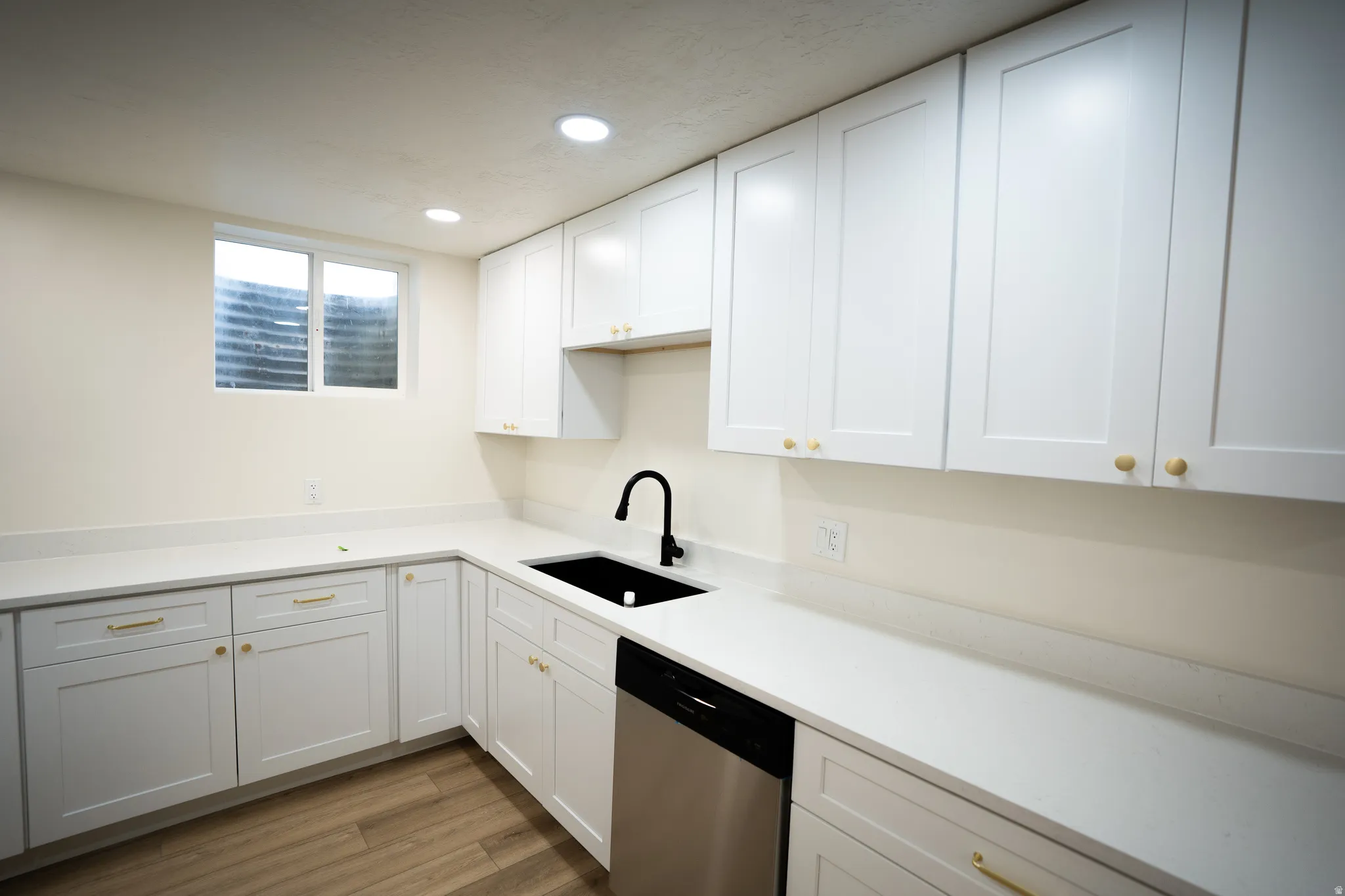 Modern kitchen featuring white shaker-style cabinetry with gold hardware, light-toned countertops, a black undermount sink with a black faucet, a stainless steel dishwasher, and recessed ceiling lighting
