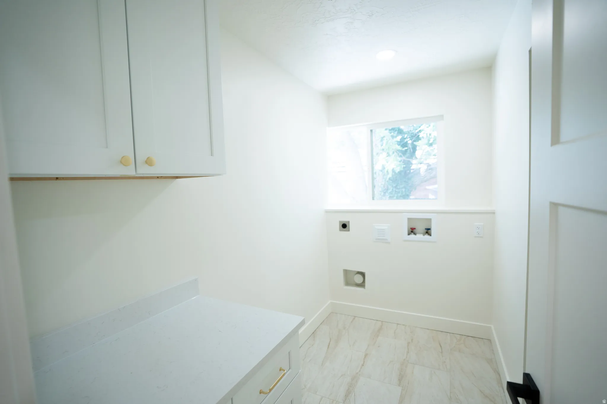 Dedicated utility room featuring light-toned tile flooring, a window, white cabinetry with gold-toned hardware, a light-toned countertop, and modern white paneled door