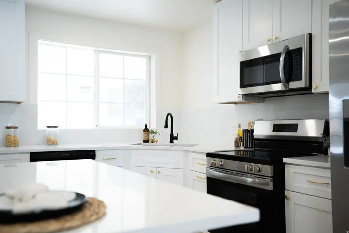 Modern kitchen featuring white shaker cabinetry, stainless steel appliances, a white solid-surface countertop, an undermount sink with a matte black gooseneck faucet, and a large window with grid panes