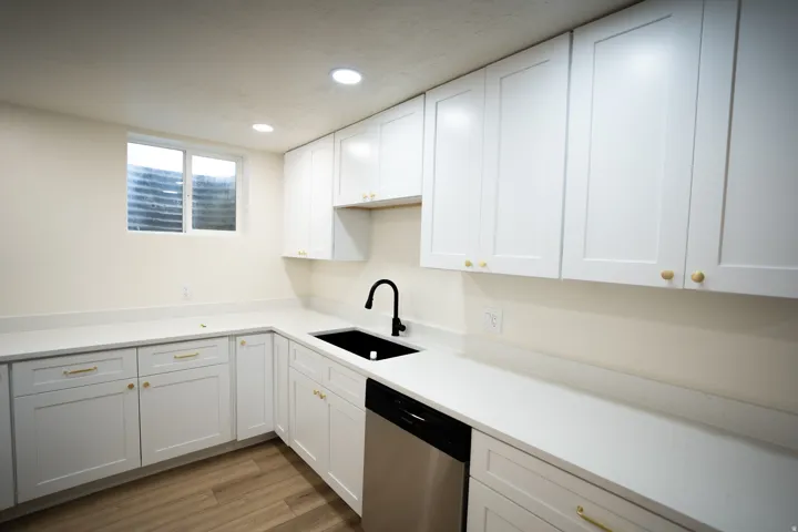 Modern kitchen featuring white shaker-style cabinetry with gold hardware, light-toned countertops, a black undermount sink with a black faucet, a stainless steel dishwasher, and recessed ceiling lighting