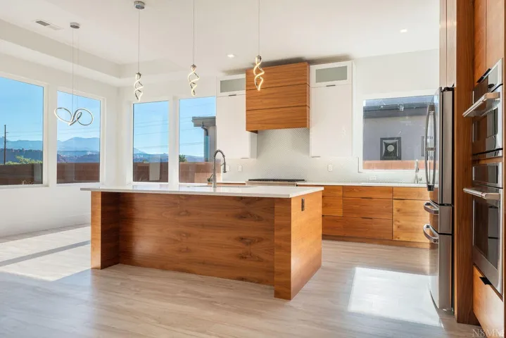 Contemporary kitchen featuring a waterfall-edge island with integrated sink, stainless steel appliances, wood-finish cabinetry, white upper cabinets, and large picture windows with mountain scenery