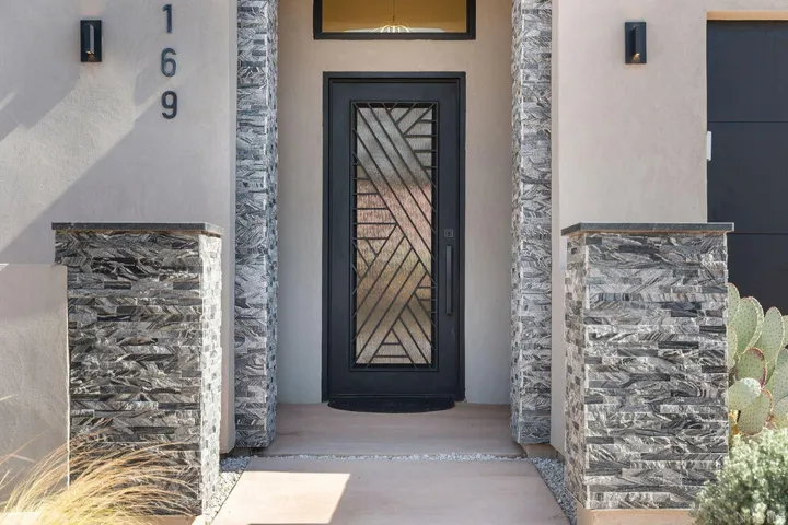 Entryway featuring a decorative wrought iron and glass door, stucco exterior, stacked stone architectural accents, and modern wall sconces