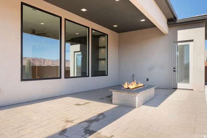 Outdoor covered patio featuring a built-in fire pit, three large black-framed windows, recessed ceiling lighting, and a paved stone surface