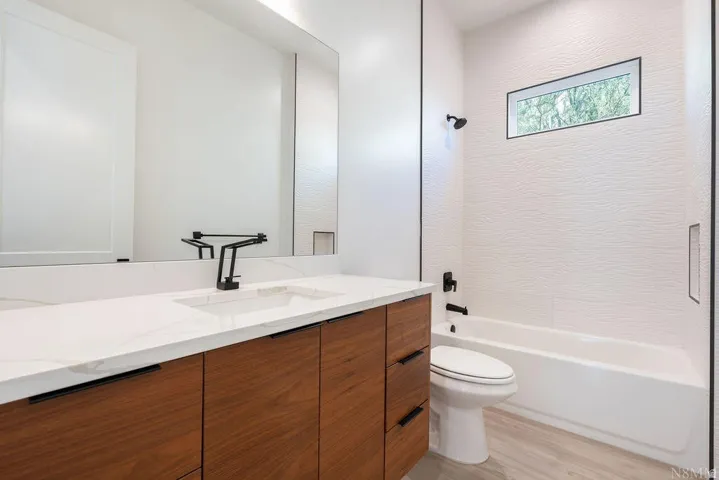 Bathroom featuring a wood-finish vanity with a white countertop and integrated sink, a large wall-mounted mirror, matte black fixtures, a white bathtub, and textured white wall tiling