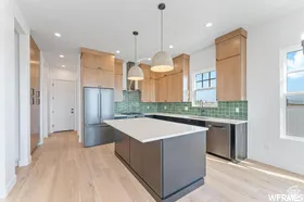 Modern kitchen featuring a central island with white countertop, light wood-finish flooring, stainless steel appliances, contemporary light wood cabinetry, and a green tiled backsplash