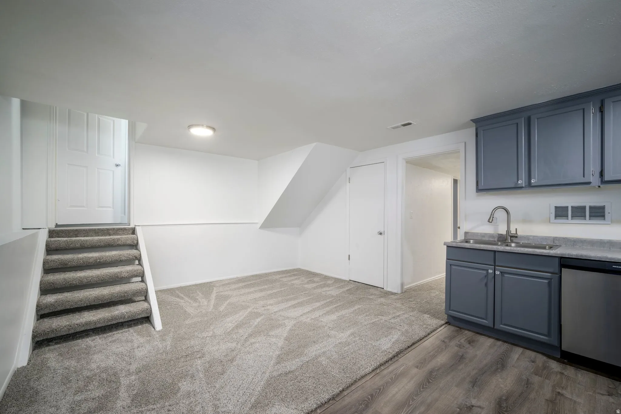 Finished basement area featuring carpeted flooring, a dedicated staircase with a privacy door, a built-in wet bar with gray cabinetry, a stainless steel sink, and wood-finish flooring