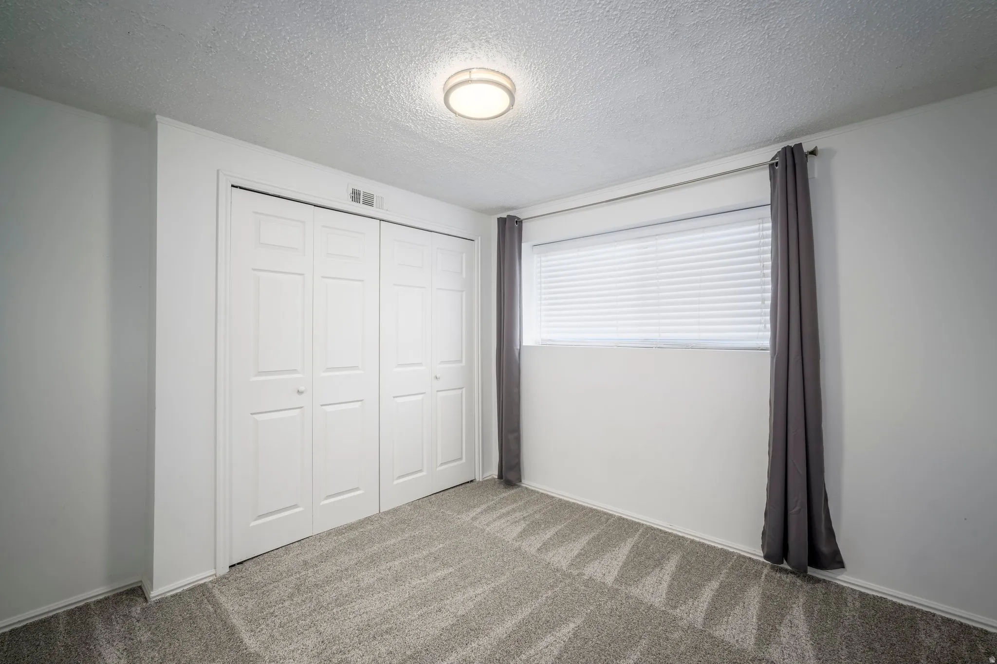 Room featuring a wide window with blinds and dark gray drapes, neutral wall paint, and patterned gray carpeting