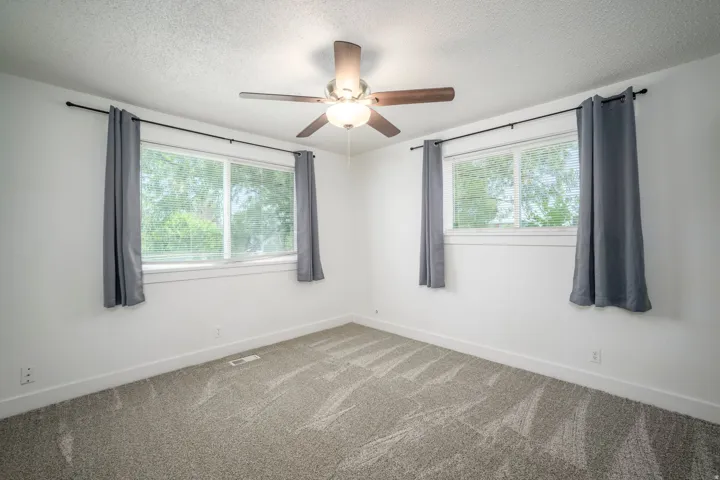 Bright room featuring two windows with white blinds, neutral carpeting, white walls, and a ceiling fan with integrated lighting