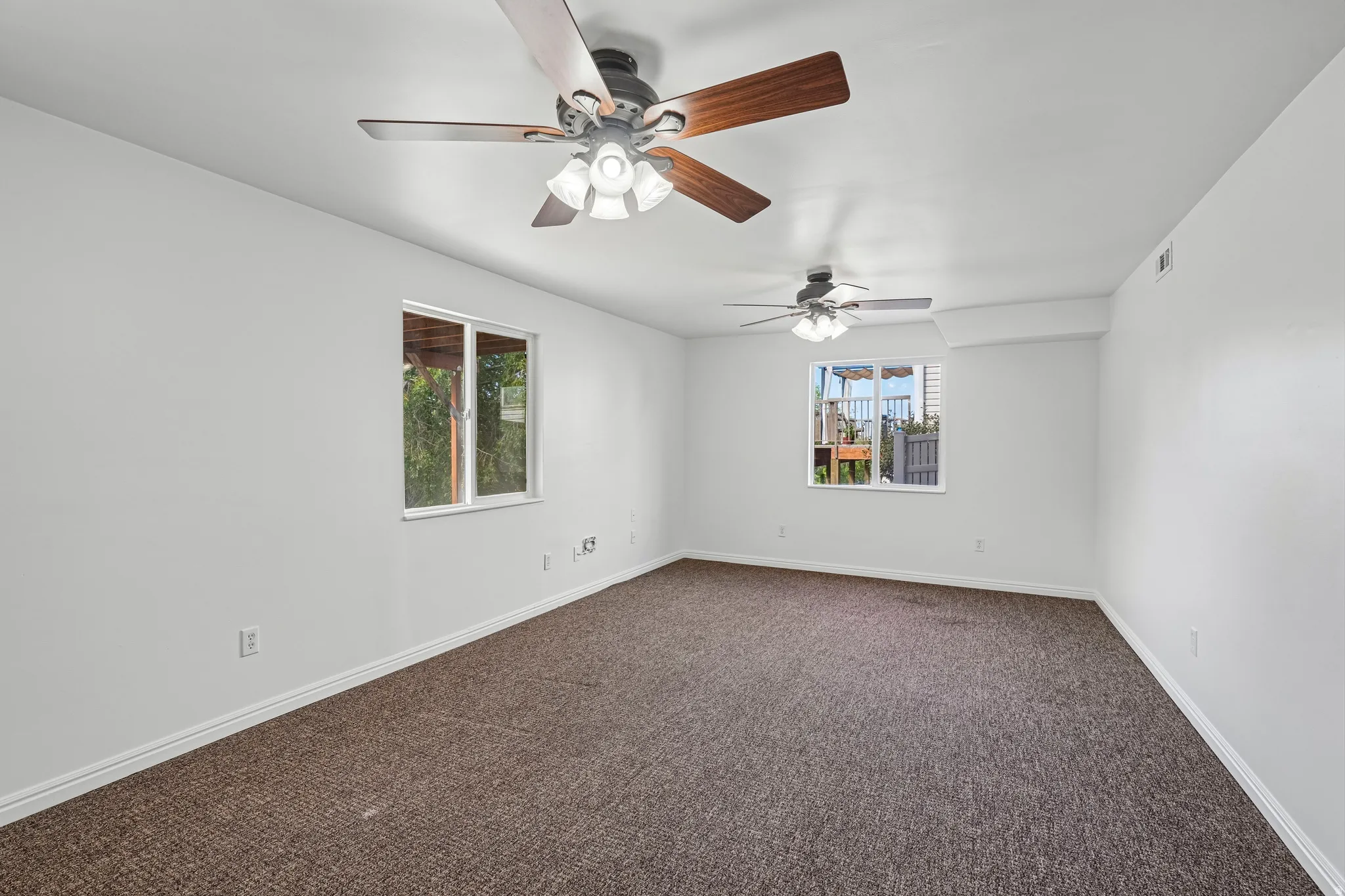 Spacious interior room featuring neutral wall tones, two ceiling fans with integrated lighting, two windows, and a textured carpeted floor