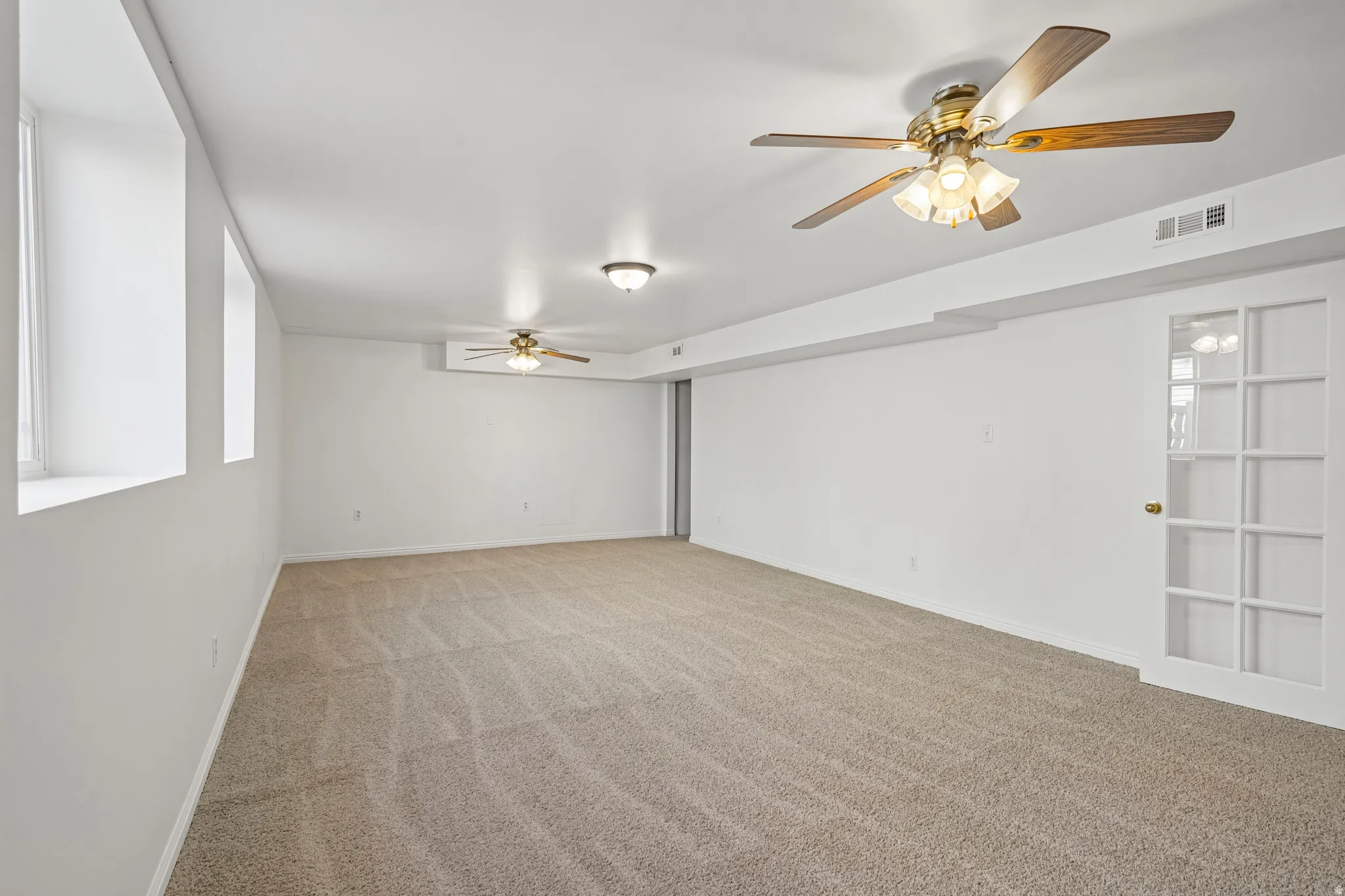 Spacious room featuring neutral carpeting, white walls, two ceiling fans with integrated lighting, and a white French door with multiple glass panes